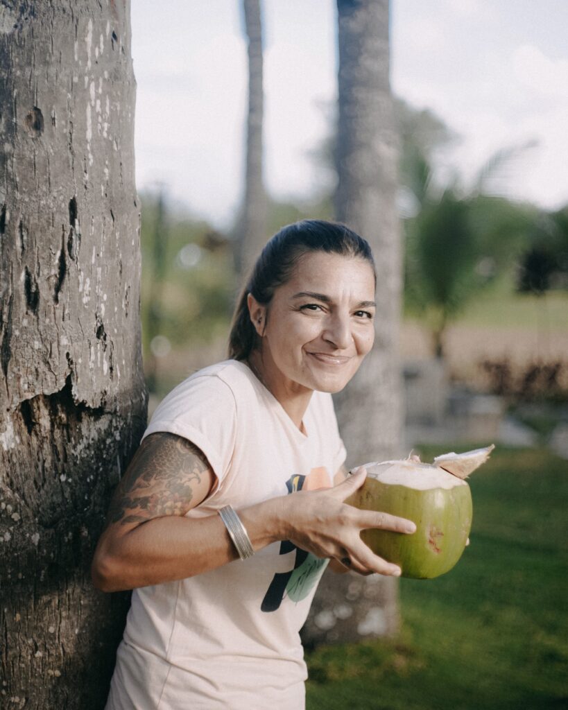Frau hält eine Kokosnuss in der Hand, lehnt lachend an einer Palme in Indonesien.